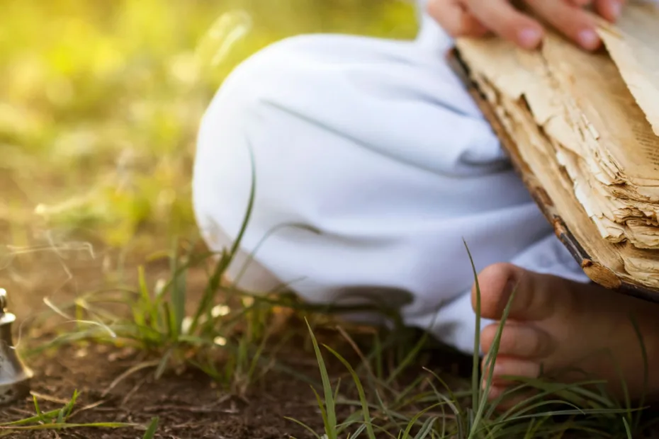 Uma pessoa sentada na grama abrindo um livro antigo ao ar livre, com sinos ao lado, sugerindo estudo ou contemplação de uma sabedoria tradicional.
