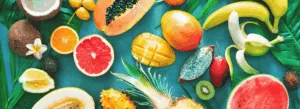 Uma variedade de frutas tropicais, incluindo mamão, banana, manga, abacaxi, coco, melancia e outras, dispostas sobre folhas verdes.