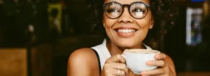 Uma mulher sorridente de óculos segura uma xícara de café em um ambiente de cafeteria.