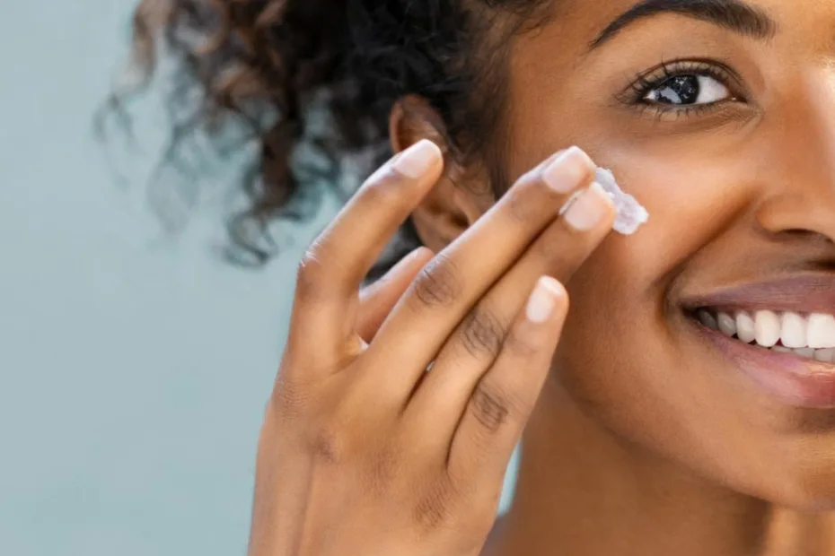 Mulher sorridente aplicando um creme na bochecha. Ela tem cabelos cacheados e uma expressão alegre, em um fundo azul suave.