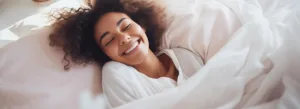 Uma mulher de cabelos cacheados está deitada na cama, sorrindo de olhos fechados, rodeada por lençóis brancos em um quarto bem iluminado.