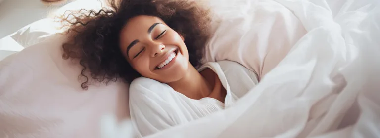 Uma mulher de cabelos cacheados está deitada na cama, sorrindo de olhos fechados, rodeada por lençóis brancos em um quarto bem iluminado.
