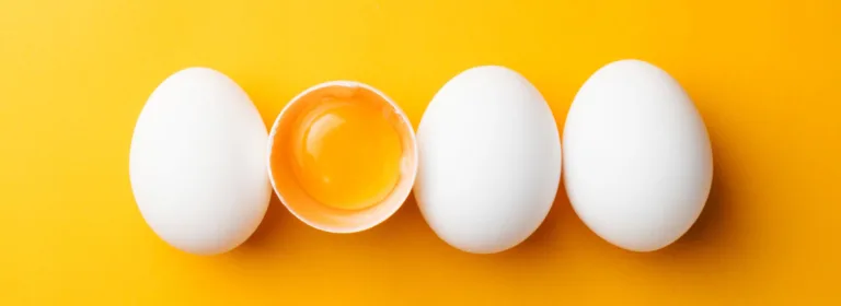 Quatro ovos brancos estão alinhados horizontalmente sobre um fundo amarelo brilhante. O segundo ovo está rachado, revelando a gema.