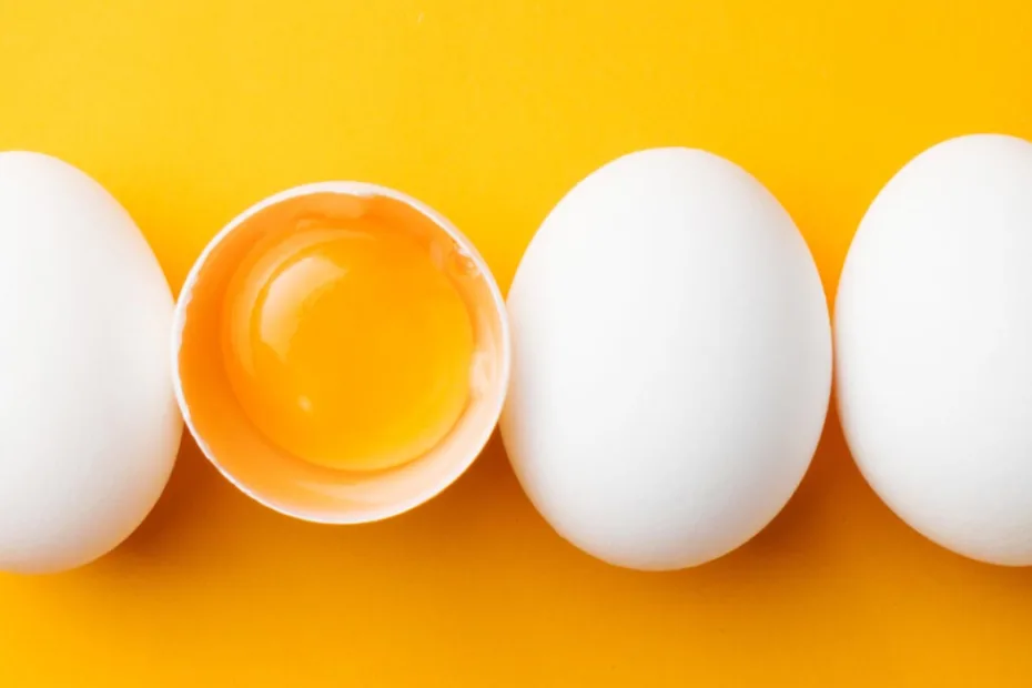 Quatro ovos brancos estão alinhados horizontalmente sobre um fundo amarelo brilhante. O segundo ovo está rachado, revelando a gema.