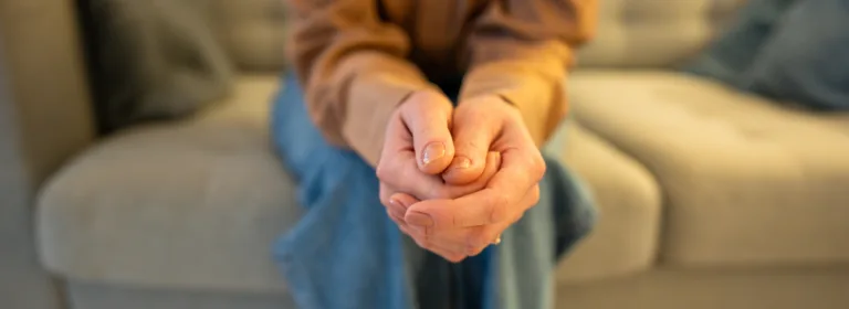 Pessoa sentada em um sofá claro, com as mãos entrelaçadas em primeiro plano, em gesto de tensão ou apreensão; veste blusa bege e calça jeans, em ambiente interno com luz quente e fundo desfocado.