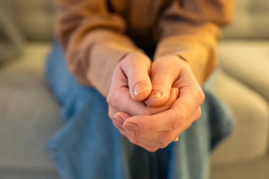 Pessoa sentada em um sofá claro, com as mãos entrelaçadas em primeiro plano, em gesto de tensão ou apreensão; veste blusa bege e calça jeans, em ambiente interno com luz quente e fundo desfocado.