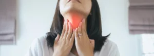 Uma mulher de camisa branca segura a garganta, destacada em vermelho, indicando dor ou desconforto.