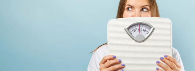 Uma mulher de cabelos castanho-claros segura uma balança branca, que cobre parcialmente seu rosto. Fundo azul-claro.