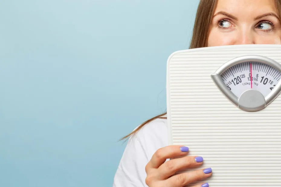 Uma mulher de cabelos castanho-claros segura uma balança branca, que cobre parcialmente seu rosto. Fundo azul-claro.