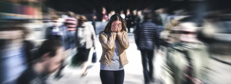 Uma mulher está de pé em uma rua movimentada e desfocada, cobrindo o rosto com as mãos, transmitindo uma sensação de ansiedade ou pânico.