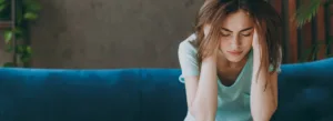 Uma mulher está sentada em um sofá azul, inclinada para a frente com a cabeça entre as mãos, parecendo estressada. O ambiente tem um fundo calmo, em tons neutros, com plantas.