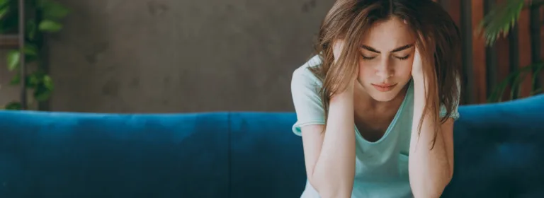 Uma mulher está sentada em um sofá azul, inclinada para a frente com a cabeça entre as mãos, parecendo estressada. O ambiente tem um fundo calmo, em tons neutros, com plantas.