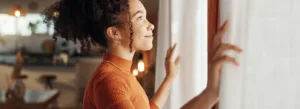 Mulher de blusa laranja abre as cortinas, sorrindo enquanto a luz do sol entra.