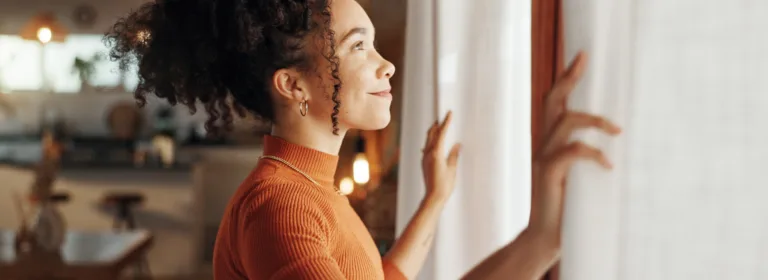Mulher de blusa laranja abre as cortinas, sorrindo enquanto a luz do sol entra.