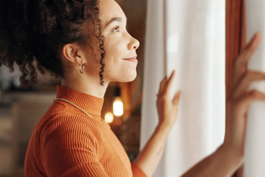 Mulher de blusa laranja abre as cortinas, sorrindo enquanto a luz do sol entra.