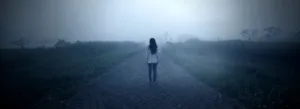 Uma figura solitária, vestindo jeans e camisa branca, está em um caminho enevoado e desolado, com grama alta em ambos os lados, evocando uma sensação de solidão e mistério.