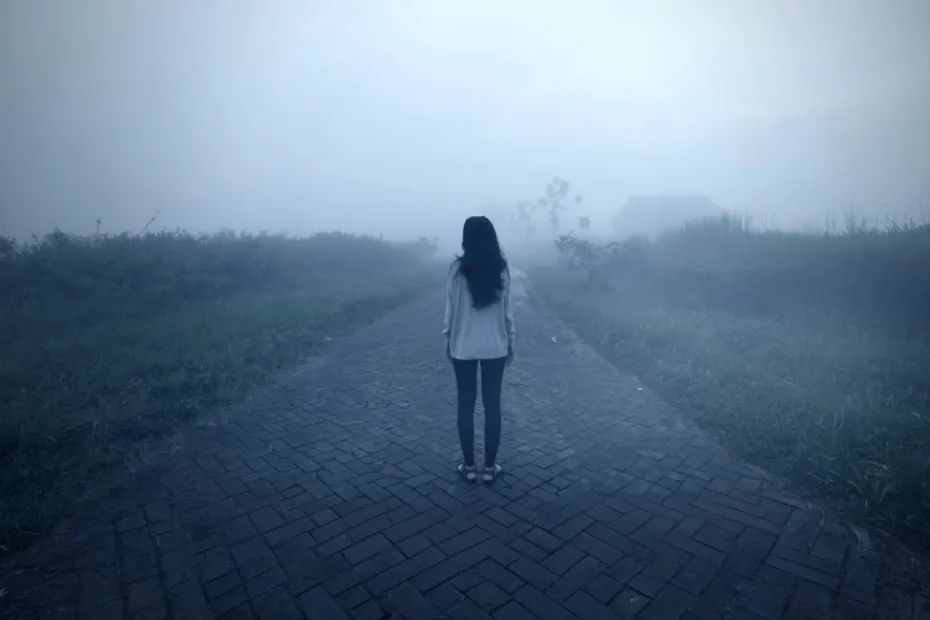 Uma figura solitária, vestindo jeans e camisa branca, está em um caminho enevoado e desolado, com grama alta em ambos os lados, evocando uma sensação de solidão e mistério.