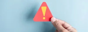 Uma mão segura um pequeno sinal de advertência triangular vermelho com um ponto de exclamação amarelo sobre um fundo azul claro, transmitindo cautela ou alerta.