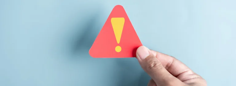 Uma mão segura um pequeno sinal de advertência triangular vermelho com um ponto de exclamação amarelo sobre um fundo azul claro, transmitindo cautela ou alerta.