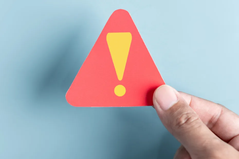 Uma mão segura um pequeno sinal de advertência triangular vermelho com um ponto de exclamação amarelo sobre um fundo azul claro, transmitindo cautela ou alerta.