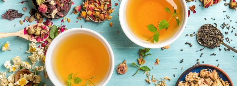 Duas xícaras de chá de ervas com folhas de hortelã sobre uma superfície de madeira azul, rodeadas por flores secas, pétalas e especiarias, criando uma cena vibrante e relaxante.