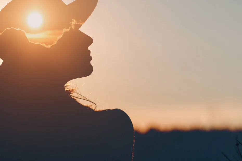 Silhueta de uma mulher de chapéu ao pôr do sol, com o sol criando um efeito de halo em sua cabeça.