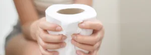 Pessoa sentada segurando um rolo de papel higiênico com as duas mãos; camisa listrada visível, rosto não mostrado.