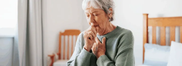 Pessoa idosa tossindo com a mão fechada próxima à boca, sentada em um ambiente claro, representando sintomas respiratórios como tosse persistente e enfraquecimento.