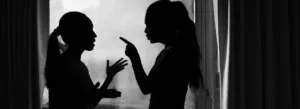 Silhuetas de duas mulheres frente a frente diante de uma janela, em aparente discussão. Uma aponta o dedo para a outra, que gesticula com as mãos abertas.